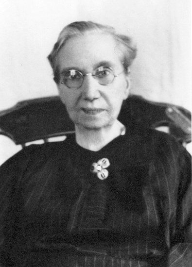 Porträttfoto (bröstbild, en face) av en äldre kvinna i glasögon, klädd
i randig blus, med en brosch framtill.