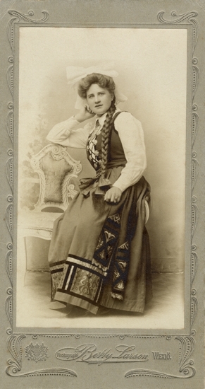 Porträtt (helfigur) av en ung kvinna i Värendsdräkt. Hon sitter på armstödet till en liten, vitmålad soffa.