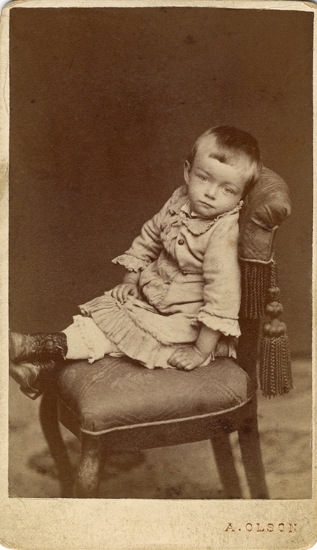 Porträtt (helfigur, halvprofil) av en liten flicka i ljus klänning med volanger, som sitter på en stoppad 
stol, prydd med fransar och tofsar.
