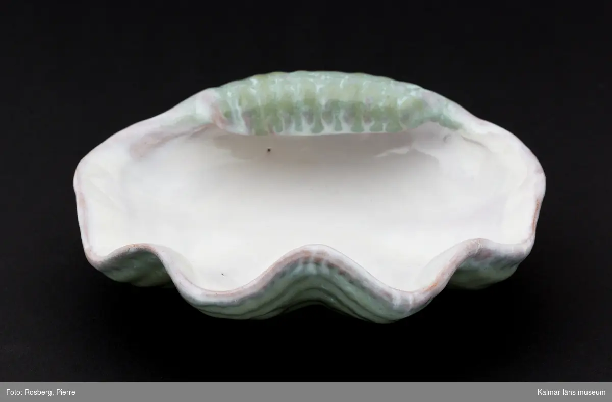KLM 45058:44. Askfat, av keramik. Askkopp av glaserad keramik i ljust grönt och rosatonat vitt. Fatet har formen av ett snäckskal. Med tillverkaretikett, tillverkat av Upsala-Ekeby.