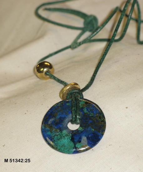 Halsband bestående av en platt cirkel av lapis lazuli i olika nyanser av blått, grönt och brunt. 
Litet hål i mitten (8 mm). Rem (900 mm) av grönfärgat läder, trädd genom hålet med ett halvslag.
En platt ring och en kula av gulmetall trädda på remmen.

Inköpt hos juvelerare Axel Ohlsson, Växjö.

Inskrivet i huvudbok 2013-11-26.