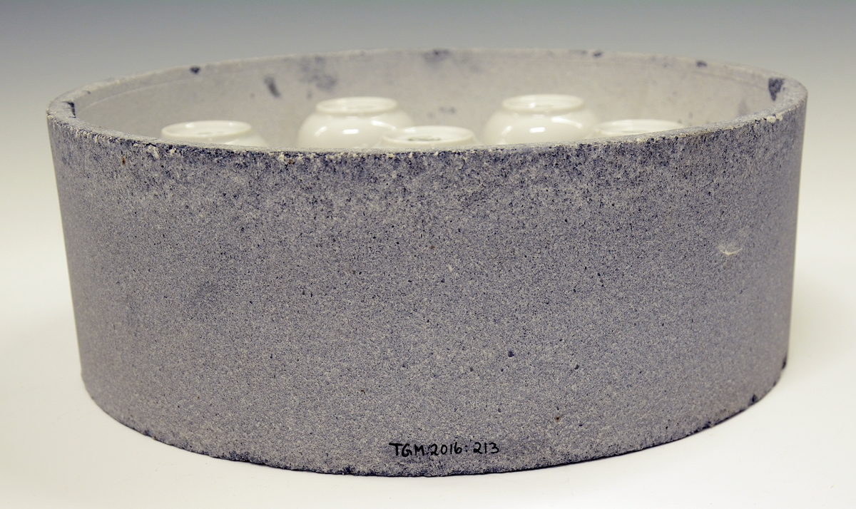 Rund kapsel i ildfast stein med syv porselenskopper på bumser/styringer (Merket A-G).