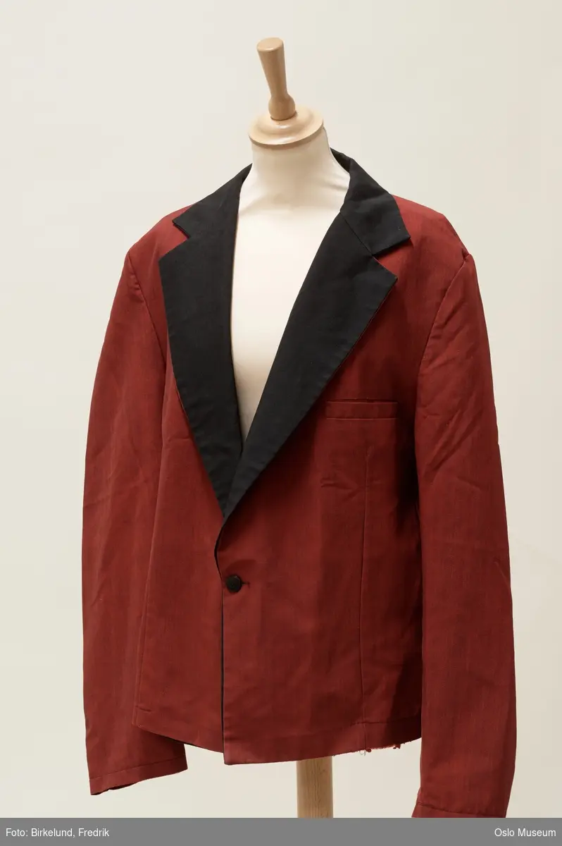 Burgunderrød jakke, kantet med sort langs hals. Sort knapp.