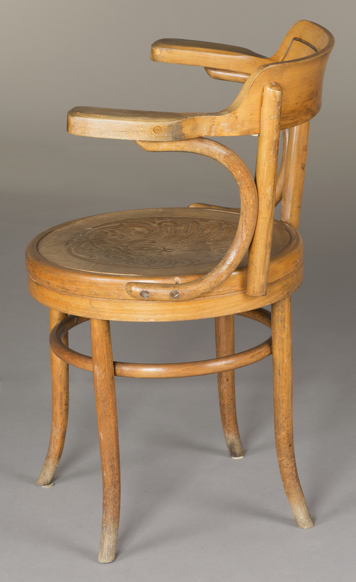 Kontorstol i lyst treverk, med armlener. Stolen har elementer av bøyd trearbeid. Et rundt svingsete. Ryggstykket har enkel linjedekor som følger formen. Setet har et utskjært mønster i form av bladverk.