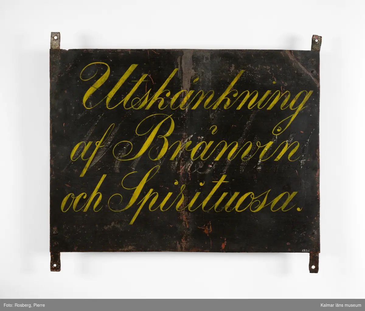 KLM 13311:1 Skylt av plåt. Svartmålad med text i gult: Utskänkning af Brännvin och Spirituosa, under denna text en äldre text: Wärdshus. Den undre texten rekonstruerad på en skylt av trä av S Wahlgren 1942, se (KLM 13311:2).