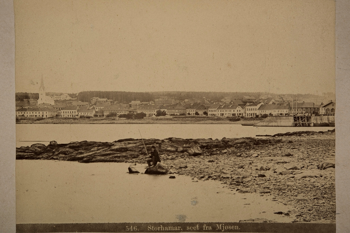 Bypanorama fra Hamar. Fisker på Tjuvholmen. Strandgata med Bekkegata og Simensgården til venstre og Jernbanebrygga til høyre. Hamarbukta. 
fotograf P. A.Thoren, Norske Prospekter. 546 "Storhamar seet fra Mjøsen"
