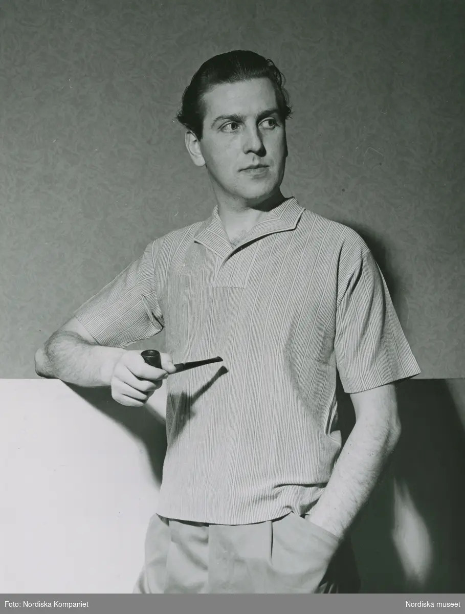 Man i smalrandig, kortärmad skjorta utan knäppning. Ljusa byxor. I ena handen håller han en pipa. Text med blyerts på baksidan: "Ital. herrmodet Italiana 1953"