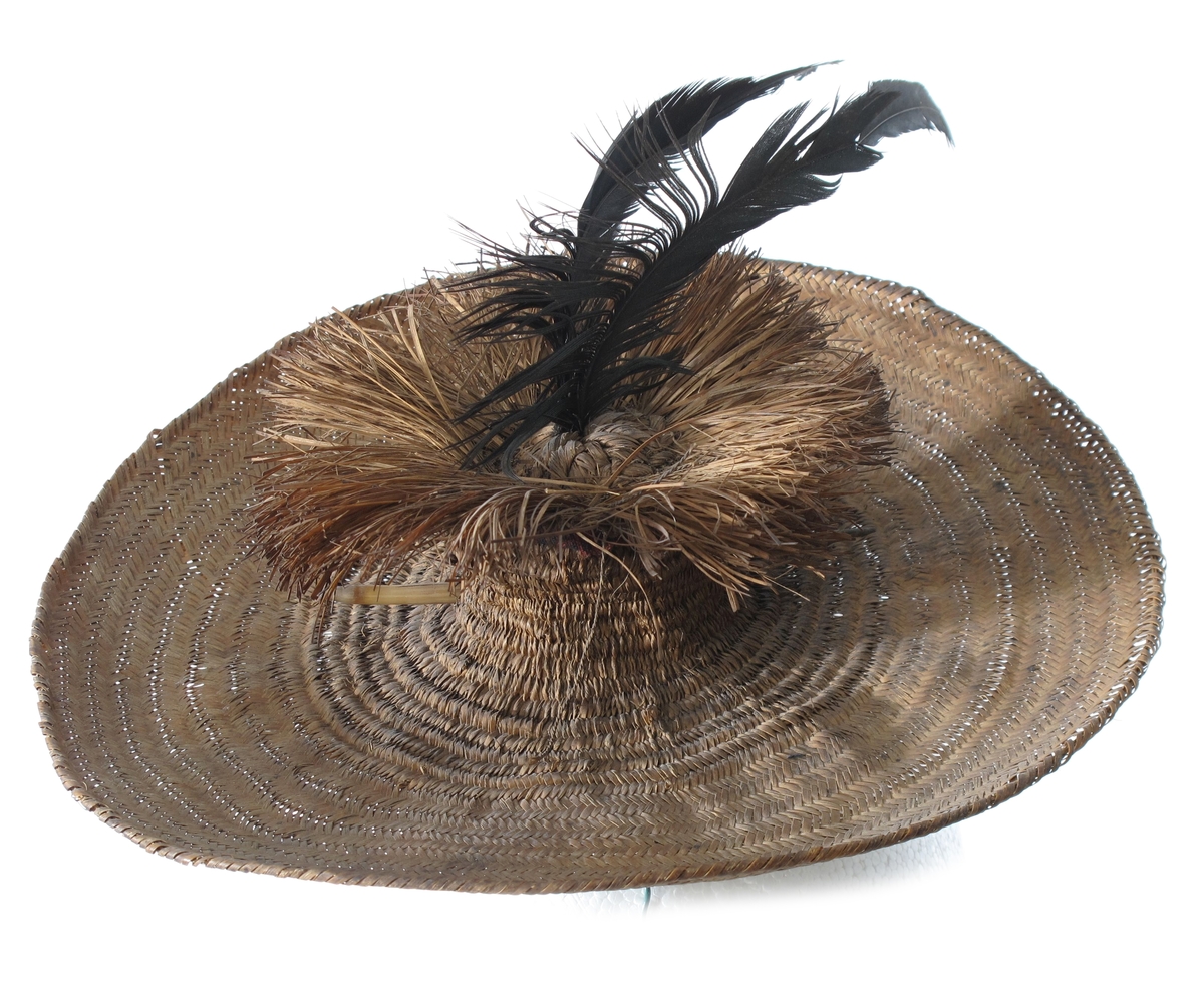 Vidbremmet hatt i mykt strå, brunt, med en liten sylindrisk  pull med  en 4,5 cm lang krans av strå rundt toppen. I toppen en sort  buet  fjær, gjennom pullen en hattenål i form av en fjærstilk.