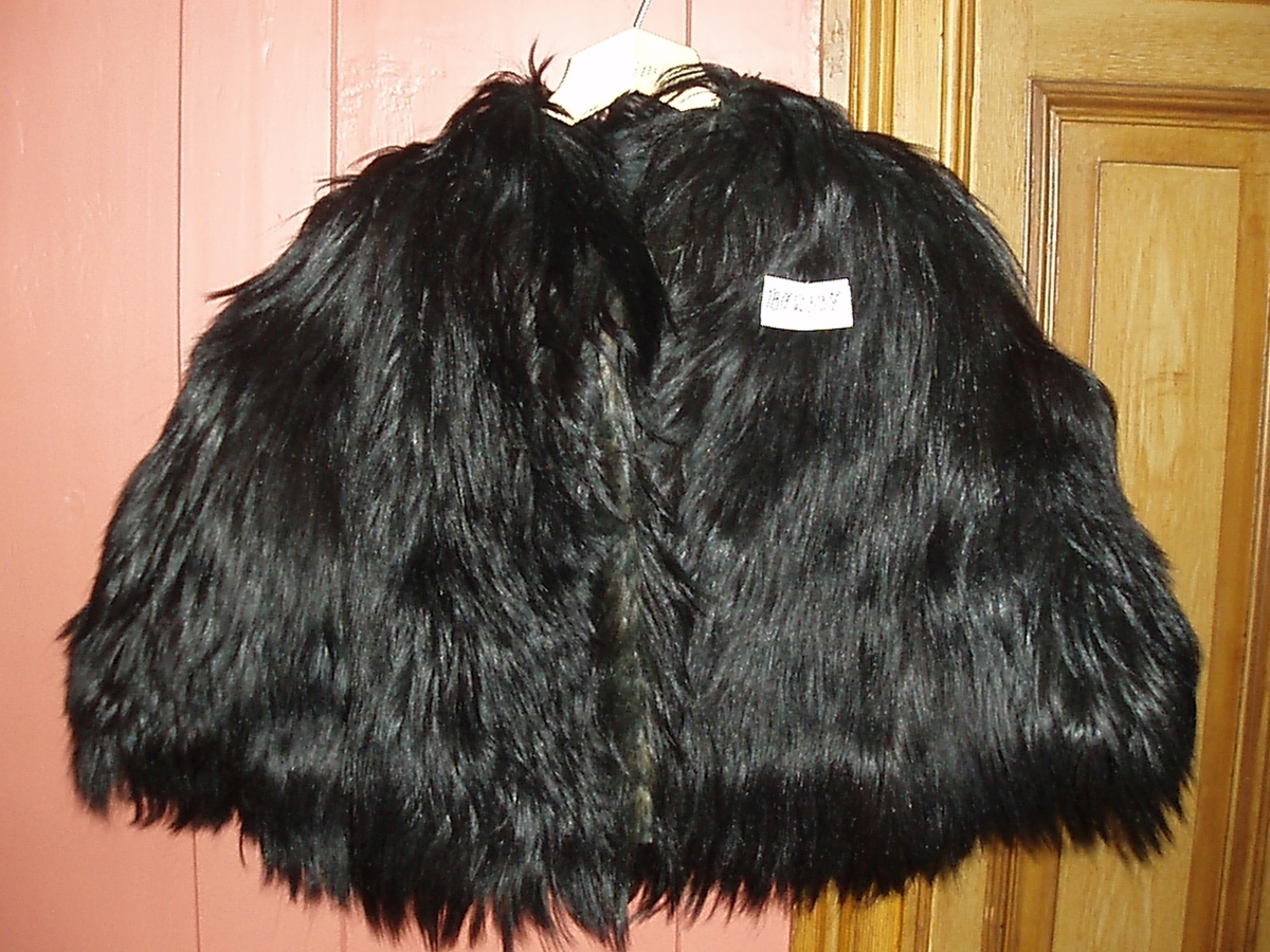 Dette er en langhåret svart pelscape. Den har vattert silkefôr, og er lukket med hekter. Inne i den er det på stramei sydd et 19,8 cm initialmerke i rødt. Merket J.Ø.S