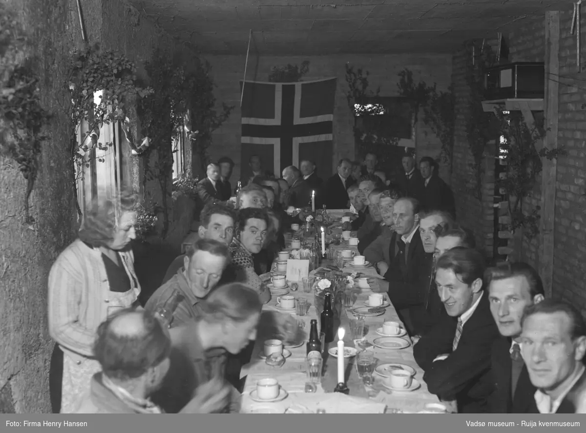 Bildet viser kranselaget på Finnmark Kringkaster i august 1948. Vi ser et langbord dekket med flasker og glass, en kvinne til venstre skjenker fra en flaske, på begge sider av bordet sitter 28 menn, mange er festkledd med dress og slips. Rommet er smalt, bordet med stolrekke på begge sider og ved kortenden dekker bordbredden rommet. På veggen til venstre er det to vinduer. Rommet er pyntet med løv og norske flagg, et stort flagg på kortveggen. På veggen til høyre står en radio på en liten hylle.
