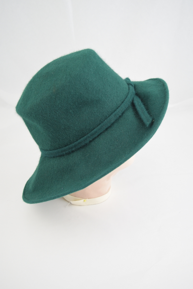 Filt, grønn, maskinsøm. Bred brem krummet opp på en side, pull sylinderformet. I overgangen mellom pull og brem er lagt et grønt bånd av samme materiale. Båndet lagt i en løkke bak og de to endestykkene er festet løst på bremmen. På det samme bånd er festet en "rosett" av svarte strutsefjær, hattens front. På innsiden av hatten er sydd et mørkere grønt hattebånd i hodeåpningen.