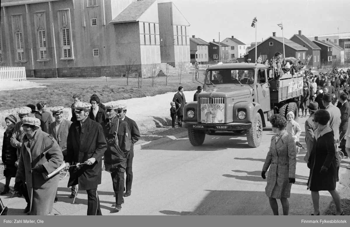 Vadsø 17.5.1969. Parade med 17.mai toget. Foran i toget sees russen, bak følger en lastebil med barnehagebarn på lasteplanet og borgertoget følger etter.  Fotoserie av Vadsø-fotografen Ole Zahl-Mölö.