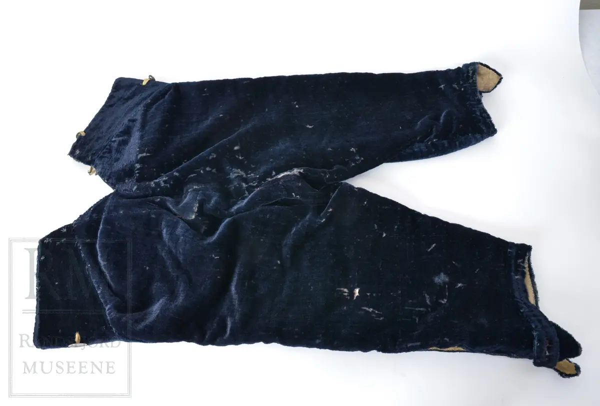 Mørk blå fløyelsbukse, knelang, med lem. Foret i lin. Fem flate messingknapper.