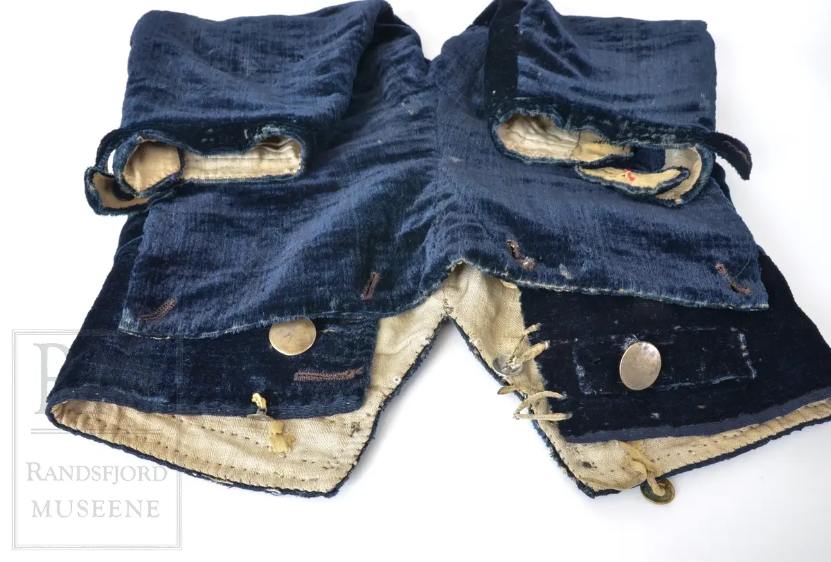 Mørk blå fløyelsbukse, knelang, med lem. Foret i lin. Fem flate messingknapper.