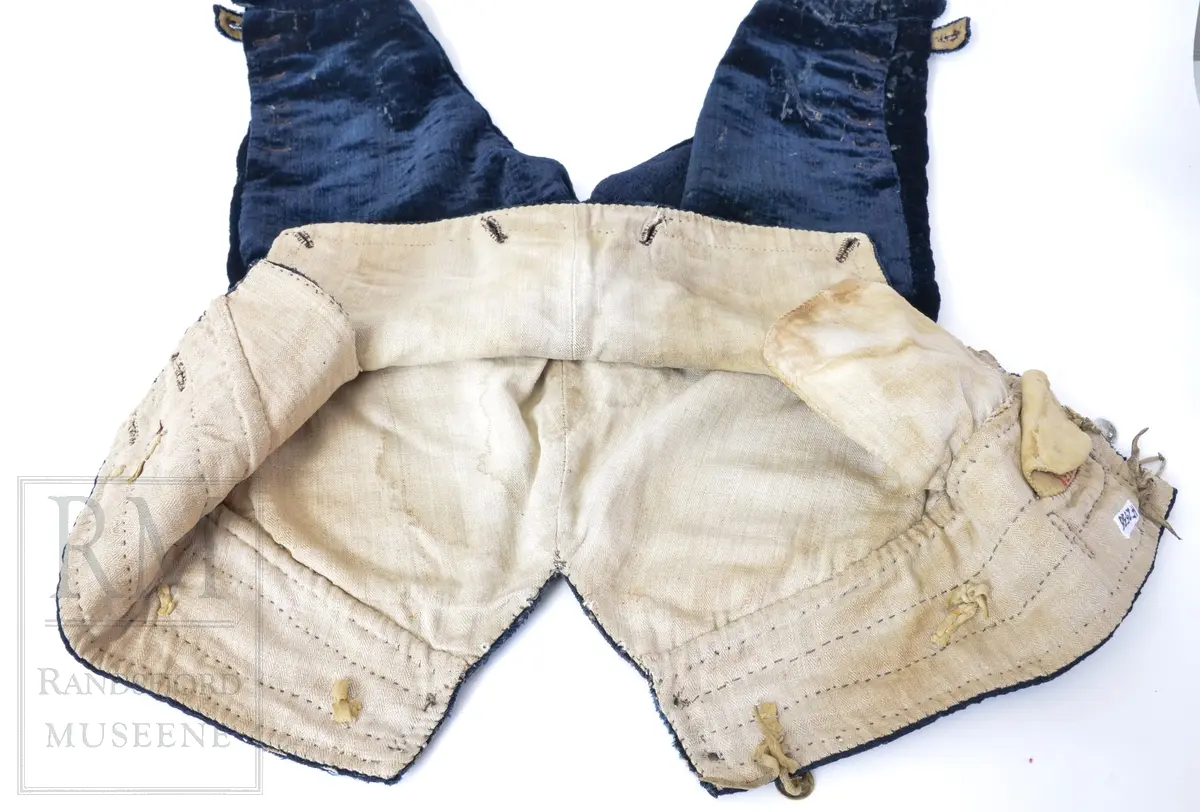 Mørk blå fløyelsbukse, knelang, med lem. Foret i lin. Fem flate messingknapper.