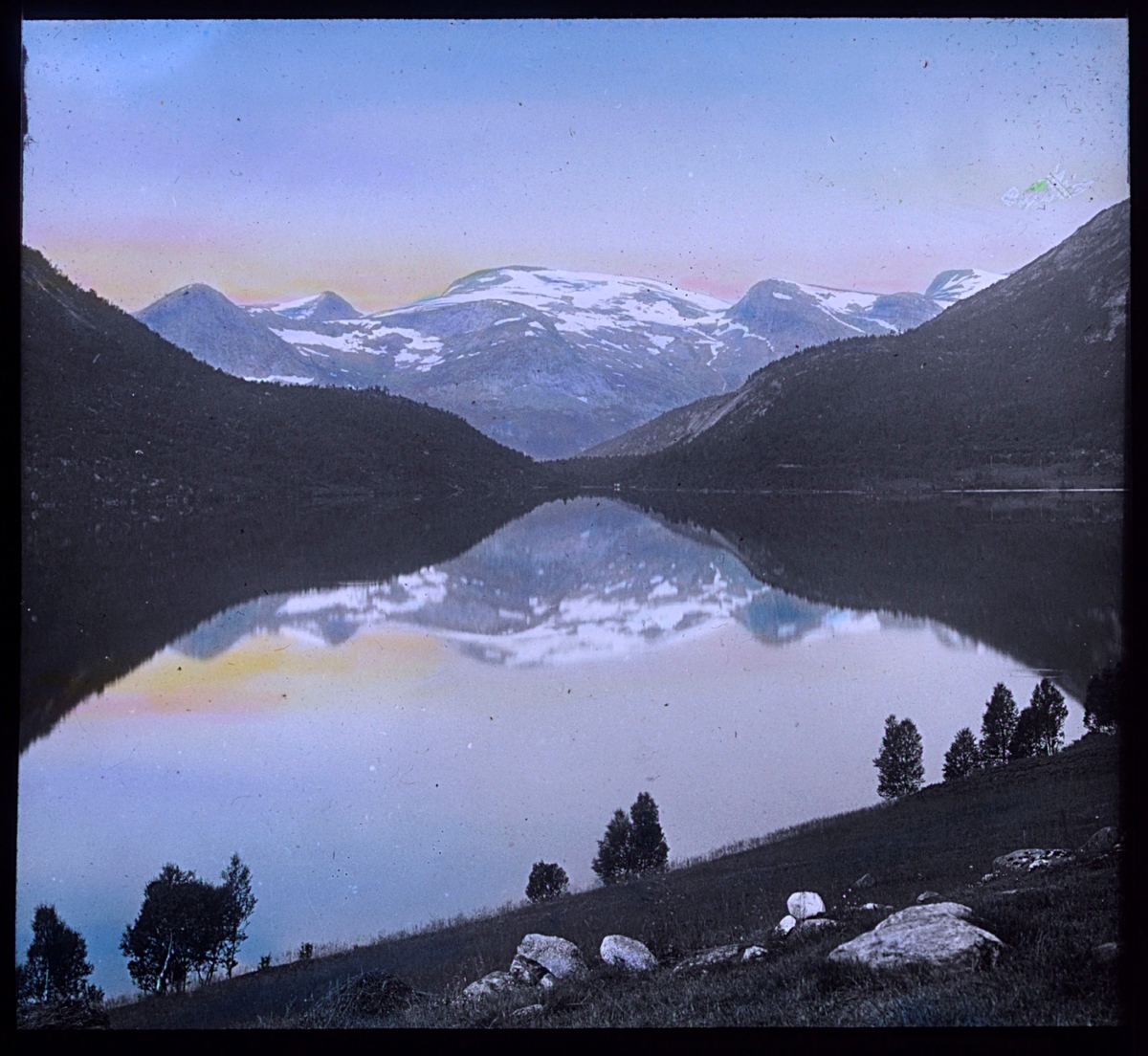 "N.842. Nordlandsnatur. N. 13. Håndkolorert av Alf Schrøder. Bildet viser et blankstille vann eller en fjord, med fjell med snø i bakgrunnen. Det er snøflekker i fjellet, men ved vannet står det bjørketrær med grønt løv. Bildet er kolorert med fargen fra sola på himmelen, muligens er det midnattsolen som er gjenskapt i pasteller med gult og rosa. Fjellet og landskapet speiler seg i vannet.