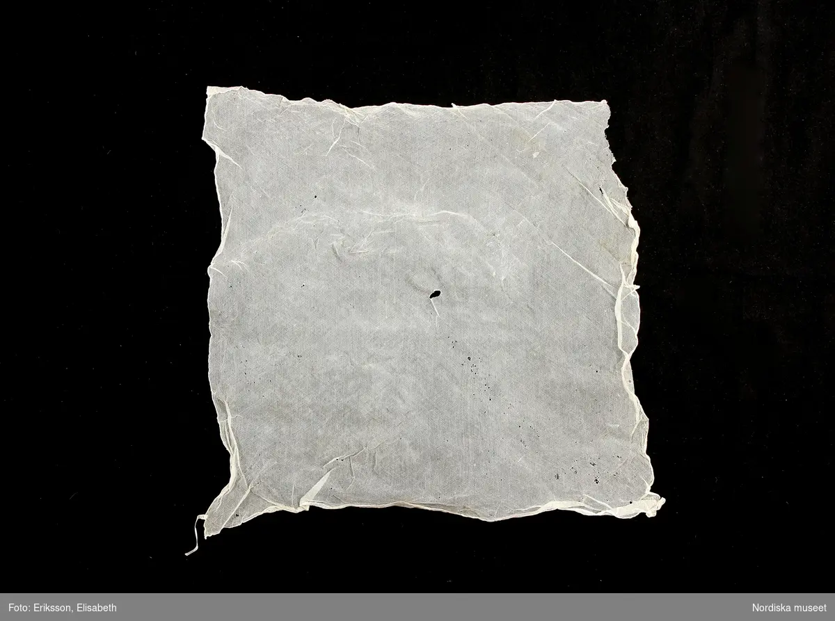 b. Kvadratiskt kläde av tunn vit  tuskaftad bomull, som batist, med smala rullfållar på alla sidor. Herlt slät, okänd användning, troligen som hals- eller huvudkläde.
/Berit Eldvik 2010-08-27