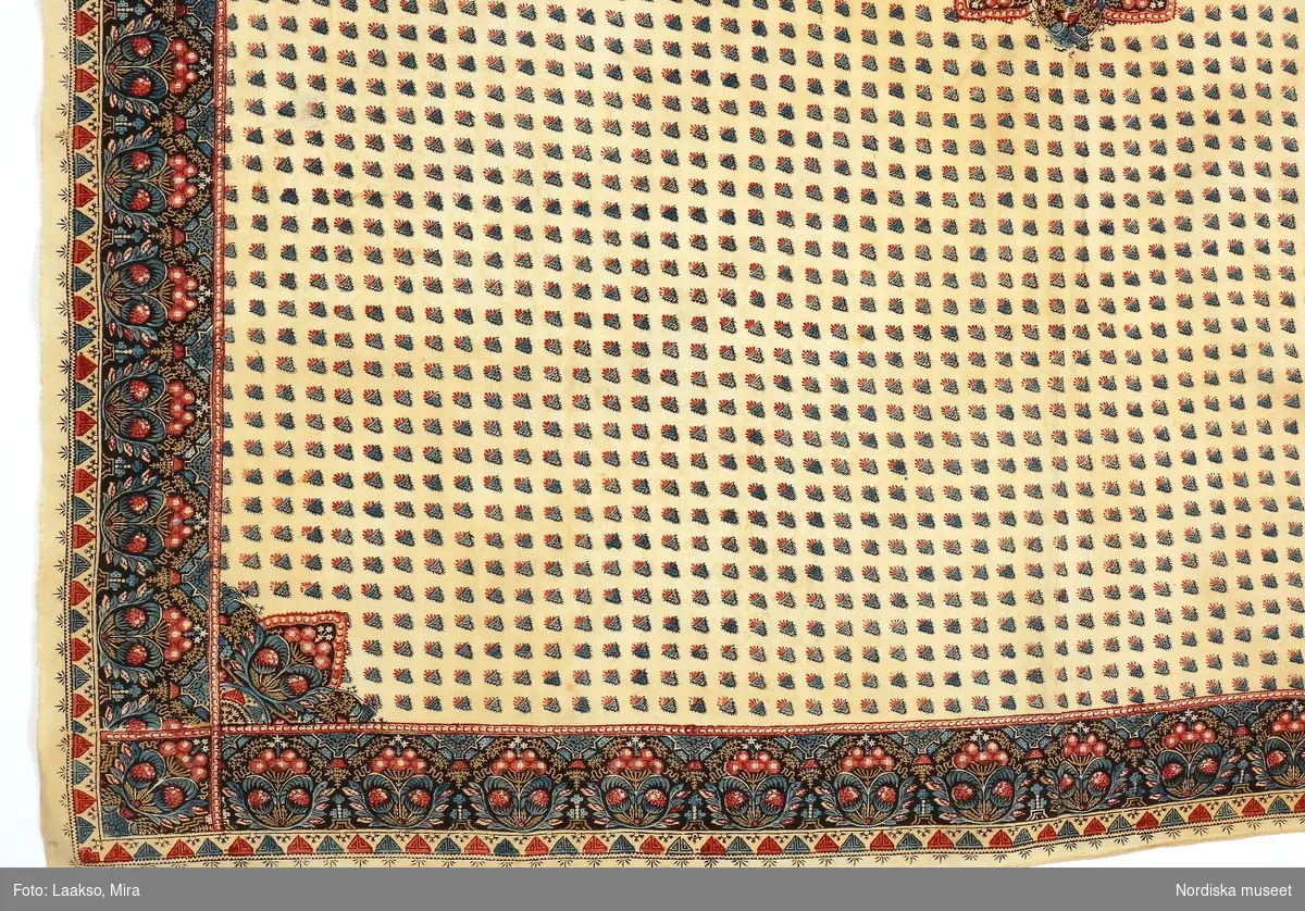 Kläde av tätvävd fintrådig bomullslärft, vit botten med handtryckt mönster som är imitation av de s.k. äkta schalarna från Kashmir. I mittspegeln ett centrumotiv med stiliserade blommor och över hela spegeln små strödda växtmotiv i grönt och rosarött. Avgränsad kantbård med i rader ställda blommotiv starkt påminnande om de äldsta miribotafigurerna i schalvävarna från Kashmir. Mönstret i rosarött, blågrönt och milt ockragult mot mörkt brun botten. Yttre smalbård med trekanter.
Trycket välgjort.
Konserverad.
/Berit Eldvik 2008-10-10
