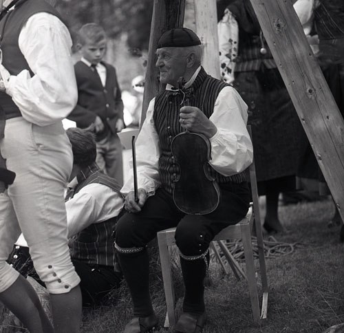 En spelman sitter med fiol och stråke i händerna 1963 i Ljusdal. Runt omkring mannen finns ytterligare människor klädda i folkdräkter.