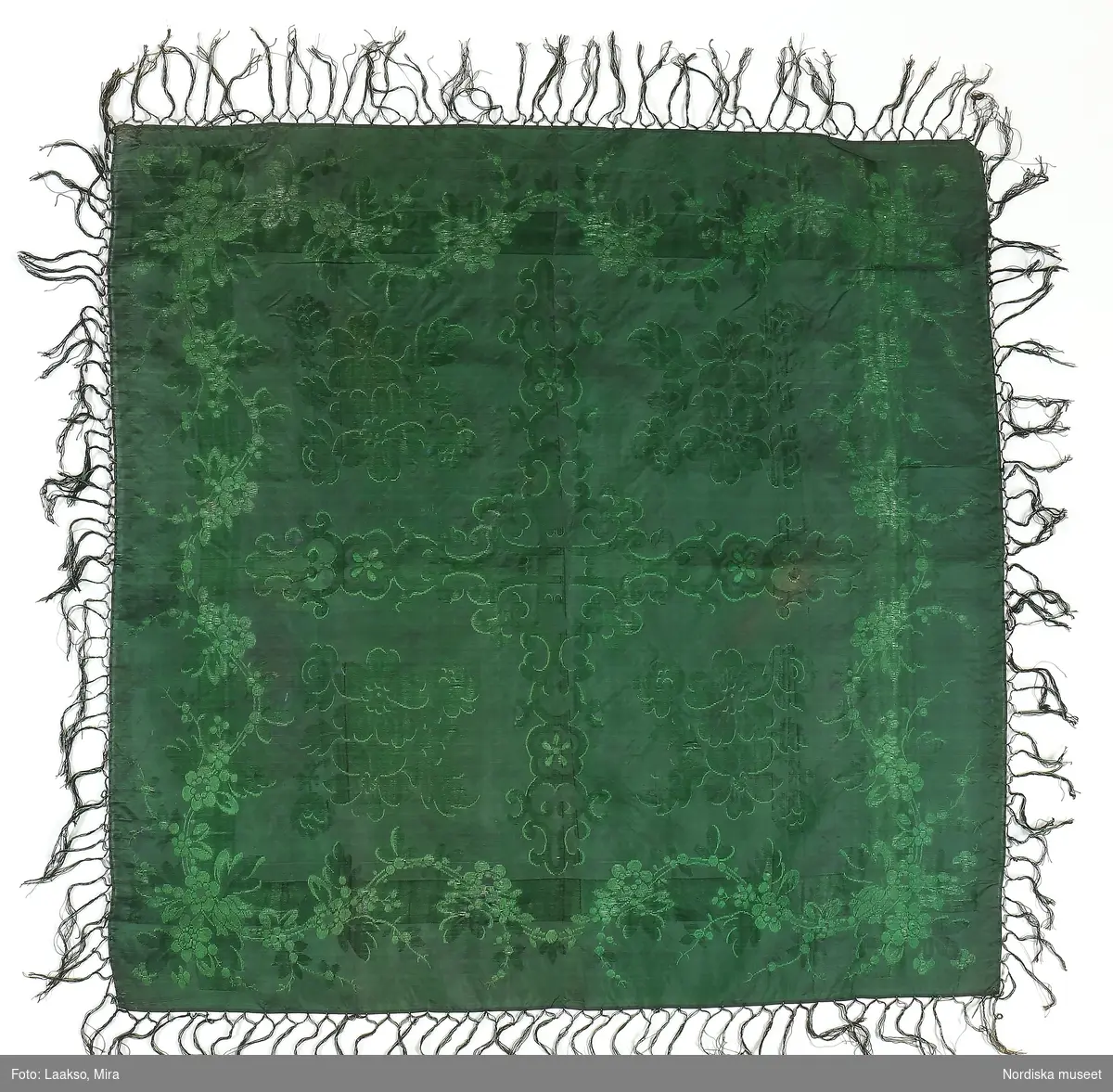 Kvadratiskt halskläde i grönsvart sidentaft med jacquardvävt mönster med inslagsflotteringar  i grönt, spegeln indelad i 4 stora rutor med ornamenterade bårder och en stor blombukett i varje ruta. Runt kanten blomslinga. Tvåknuten frans av tvinnat silke i 2 gröna nyanser.
Likadant halskläde i andra färger se 148 963 från Ilsbo, som är hallstämplat i Stockholm
/Berit Eldvik 2011-11-10