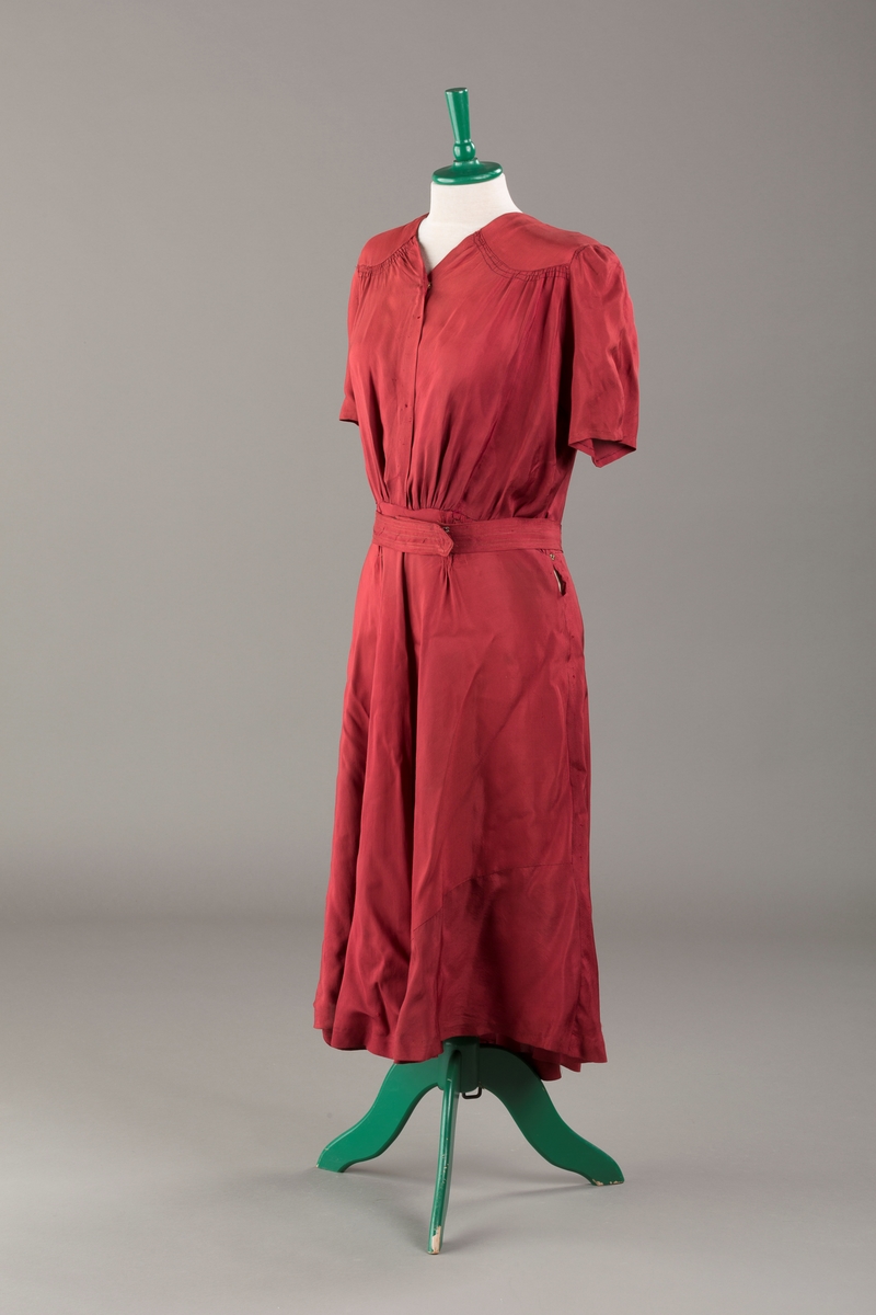 Form: Glatt, klokkeformet skjørt. Forstykkene rynket til buede bærestykker foran. Overdel rynket til skjørt. Korte ermer. Lukket med trykknapper. Belte fastsydd.