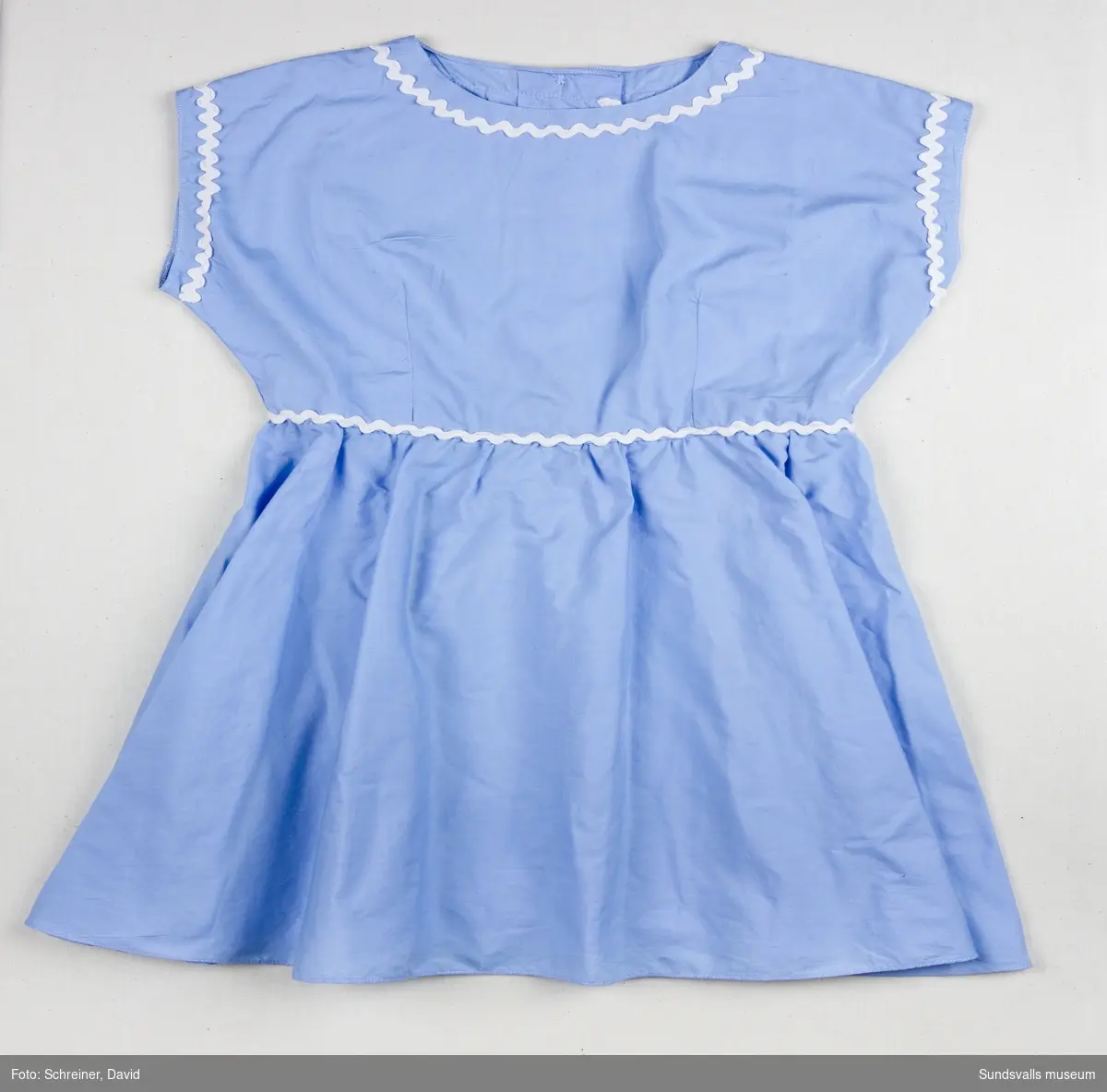 En ljusblå ärmlös klänning med utsmyckning runt ärmhål, halsrundning och midja med vitt dekorband.
