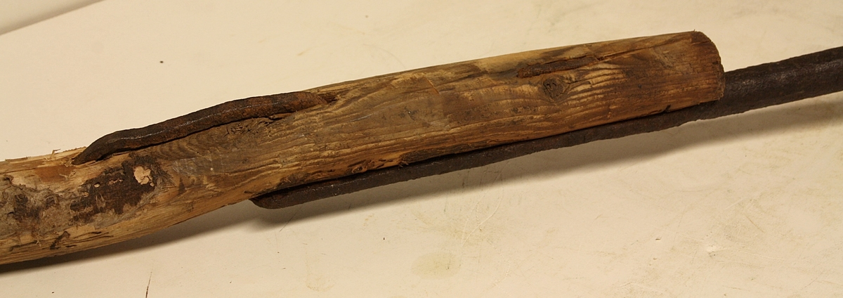 Grovt tilvirket treskaft. Langt metallskaft festet med to store spiker/nagler til treskaftet. Vifteformet, trekantet blad.