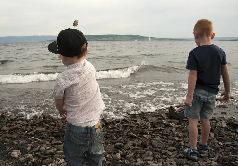 Two young boys dressed in shorts and T-shirts, one with a cap on his head, stand on a pebbly beach facing towards the water and throwing stones into it.