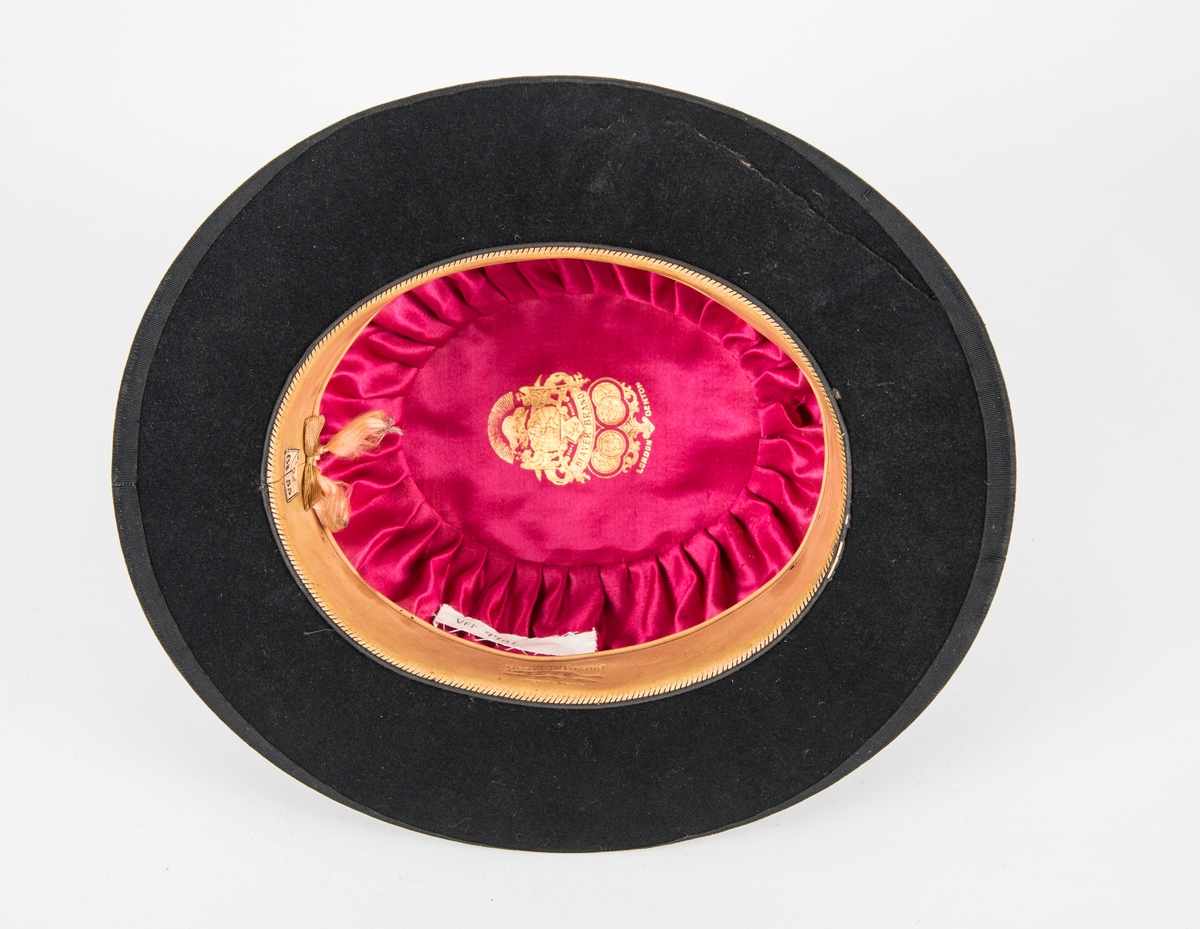 Hatt, rundpullhatt i svart filt. Ripsband rundt pullen og bremkanten. Skinnband på innsida. Fóra med rosa silke.