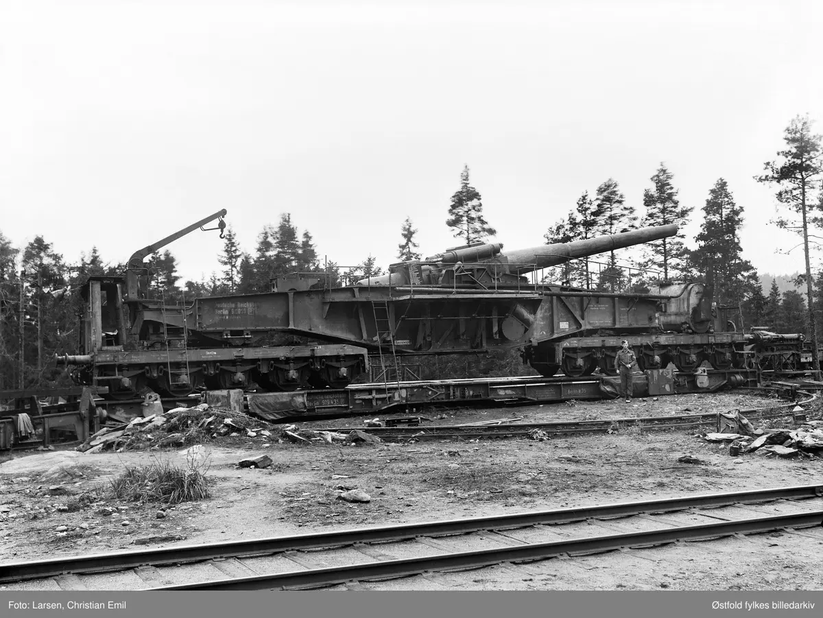 Kanon på jernbanevogn, batteriet på Knatterud, Varteig ved Sarpsborg 1945. Soldat, ukjent.
Deutsche Reichsbahn, Berlin.
E-postaddresse: gunnkull@hotmail.com

Dette er en "28 cm kanon Schwere Bruno" Den hadde en totallengde på 22.8 meter og en vekt på 118 tonn. Lengden på kanonrøret 11.93 meter. 
Utgangshastighet på granaten var 860 m/sek. Maks skuddvidde 37.8 km. Skuddhastighet ca 10 skudd i timen. Kanonbatteriet deltok først under det tyske angrepet på Frankrike i 1940, og i mars 1941 gikk det fra Sassnitz i Tyskland til Trelleborg med ferge.Deretter gjennom Sverige via Kornsjø opp til Storlien i Trøndelag. Så inn i Sverige igjen og opp til Kiruna og videre til Narvik. Jernbanekanonene ble transportert gjennom Sverige under benevnelsen "maetrialer for brubygging" Tyskerne hadde laget en ramme av vinkeljern over mekanismen og kanonløpet. Det hele ble dekket med presenning under transporten. Siden svært få hadde sett noe lignende noen gang, gikk transporten visstnok uten noen problemer. Så ble batteriet montert og var klar til bruk  på Framnesodden ved Narvik etter kort tid. I august 1942 ble batteriet beordret til Nypan ved Trondheim, også denne gang gjennom Sverige. Allerede i september samme år ble det beordret videre til Vestlandet. Der hadde det forskjellige standplasser, men fra desember 1942 ble det fast standplass ved Øyslebø, som ligger 2 mil nord for Mandal. Grunnen til at det ikke kom lenger vestover, var at jernbanen videre var smalsporet. Der ble Batterie 689 stående til januar/februar 1944 hvoretter det ble beordret til Østfold. Etter krigen sto kanonene på Mysen og det ble foretatt noe vedligehold på kanonene i 1950. Deretter, antagelig 1953-54 ble batteriet levert til Christiania Spigerverk.  Kilde: Østfold Historielags tidsskrift WIWAR nr1 2002.