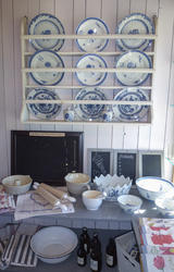 Tallerkenhylle som viser dype og flate tallerkener i hvitt porselen med blå blomsterdekor, kjøkkenhåndklær, boller, kjevler og tavler til å skrive på.