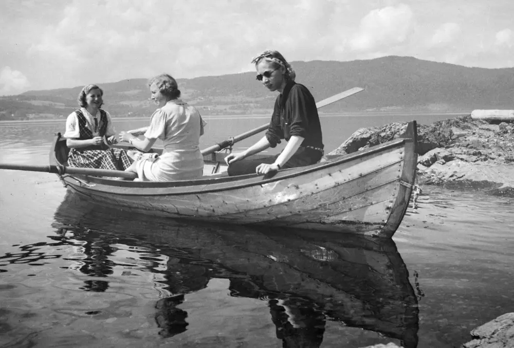 Robåt (dreggebåt, mjøsbåt, Hollandbåt), Til høyre sitter Lumi Johannessen to ukjente, utenfor "Lia, " hytta til Baker Johs. Johannessen på Jessnes i Furnes. Mjøsa. Båten er ikke malt(ant. lakkert), har utliggere for årene, typiske for mange av mjøsbåtene.