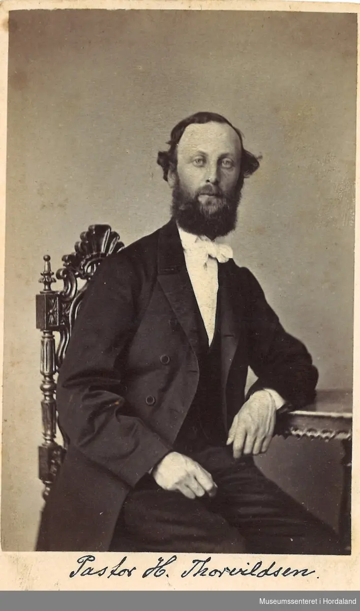 fotografi av ung mann med skjegg, svart frakk, vest og bukse, kvit skjorte med sløyfe som sit på stol