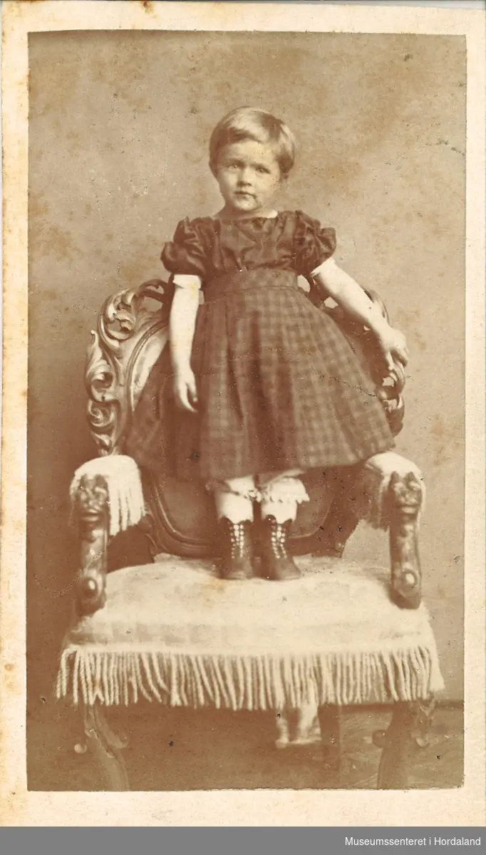 atelierfotografi av lita jente med ruta kjole, blondebukser og knappestøvlar som står på ein stol