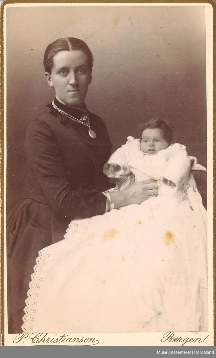 atelierfotografi av mor kledd i mørk kjole med medaljong om halsen, med barn i dåpskjole (?) på fanget