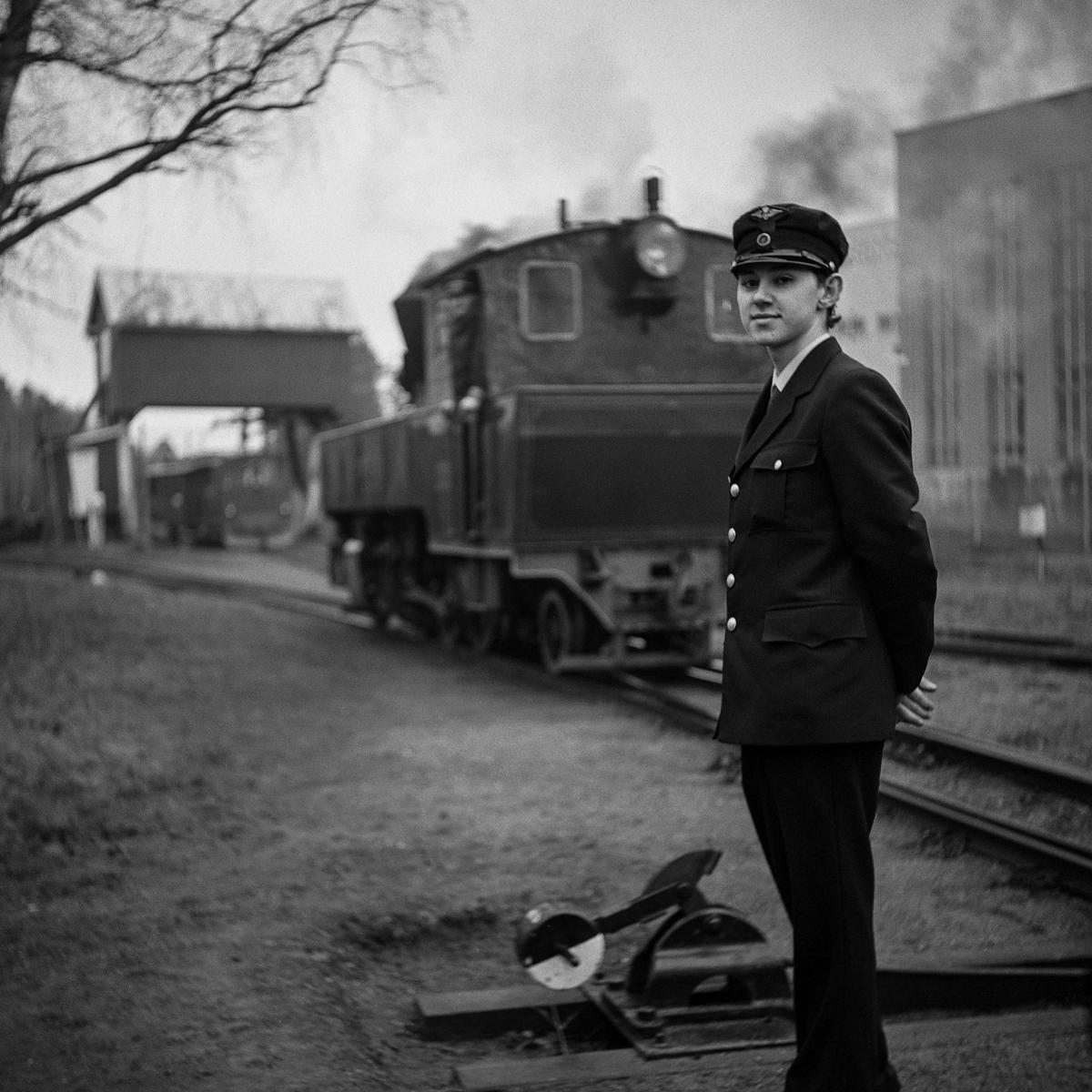 Ung mannlig frivillig i uniform foran et damplokomotiv på Sørumsand stasjon. I bakgrunnen skimtes den fredede portalkrana.