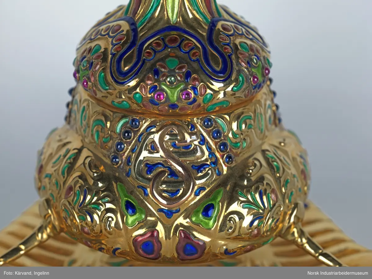Blekkhus i gull - 750 (18 karat) - med rike former og sisselert. Står på tre ben og er dekorert med ulike stener som muligens er safirer, rubiner, smaragder, perler, citriner og sølv.

Dimensjon: 17 x  17 cm
