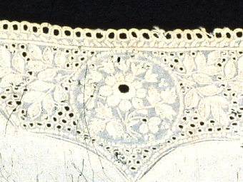 Manschett av gulvitt linne med broderi i nederkanten. Tre handsydda knapphål i en sida. Märken efter tre knappar i den andra sidan.