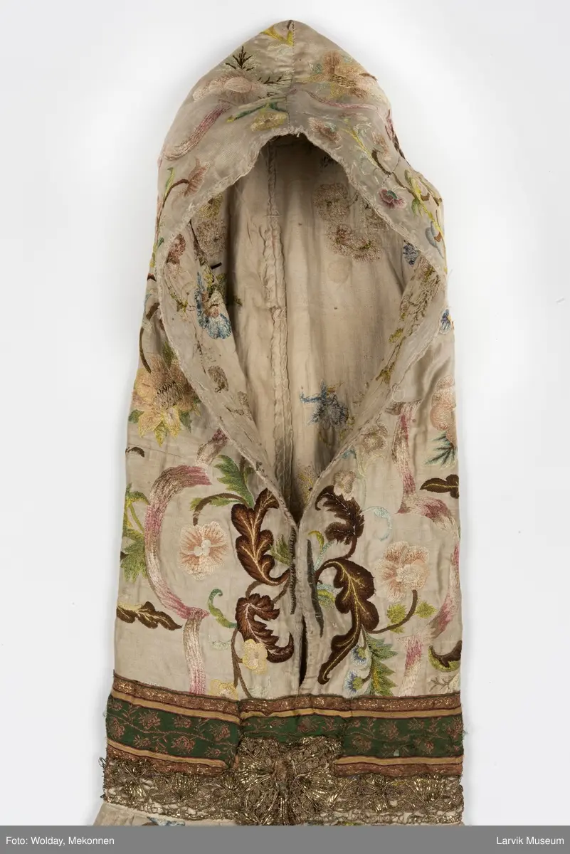 Form: livstykke som hette, åpen til livet, bred linning, rynket skjørt med blondekant nederst, søm bak på hetten i linning, søm foran og bak i skjørt, enkelte skjøtesømmer, linning brodert med gulltråd, skjørtet er foret med tynt stoff