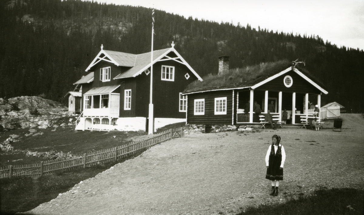 Jente i bunad, Margit (datter til Johan Galaasen, Jons (28/1 1867) stående foran hus; bevertningssted el. lign (?) v/Gausdal (?)