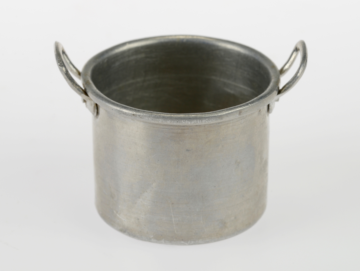 En liten komfyr med tre kasseroller og en steikepanne. Gjenstandene er undernummerert 01-05.
FTT.54454.01: En svartlakkert lekekomfyr som er firkantet med avrundede hjørner. Den er trolig laget av jernblikk og blikk. På kokeflaten er det tre hull for å plassere kasserollene på. Bakerst til venstre er det en 8cm høy pipe. I front er det en sølvfarget blikkdør som er festet med hengsler. På døren er det innskrift og et lite messingfarget og rundt håndtak. Komfyren har buede bein.
FTT.54454.02: En høy kasserolle laget av blikk. I hver side er det loddet på håndtak. 
FTT.54454.03: En lav kasserolle laget av blikk. I hver side er det loddet på håndtak.
FTT.54454.04: En lav kasserolle laget av blikk. Det har vært festet et langt håndtak i den ene siden, men det er nå løs.
FTT.54454.05: En steikepanne laget aav blikk. Håndtaket mangler.