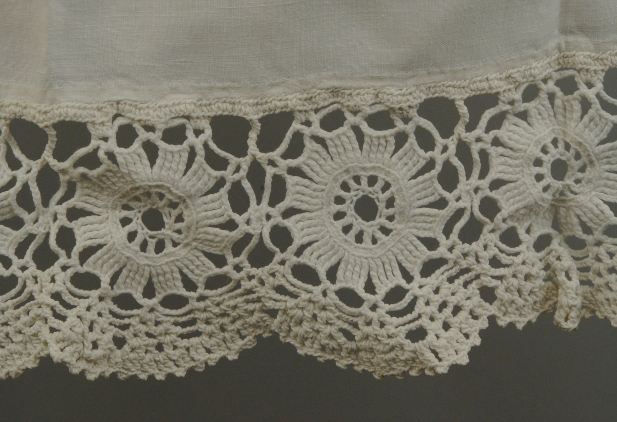 Forkle går fra livet og ned. I øvre kanten er det sydd en løpegang for bånd, ca. 1 cm brei. Ei tvunnet bomullssnor med dusker i endene er tredd inni løpegangen. Nedentil er stoffet faldet for hånd og påsydd ein håndhekla blonde i tykt bomullsgarn, det består av 10 sirkler med blonde rundt. Blonden er tunget nedentil.