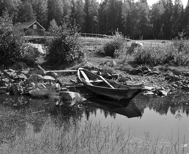Svart-hvitt foto av en tom åfløy (robåt) som ligger dratt på land med årene oppi. Stille vann og kulturlandskap med gresslette og et hus før en rekke med skog.