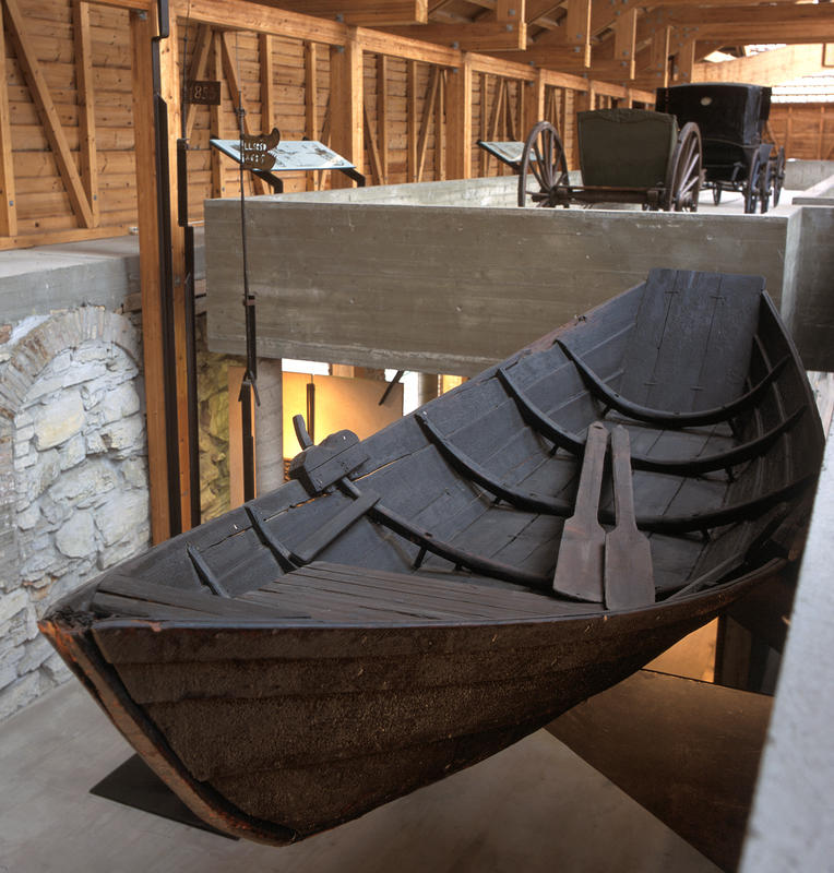 Mørkebrun robåt i tre henger oppe i utstilling i Storhamalåven, et par årer hviler i bunnen av båten.
