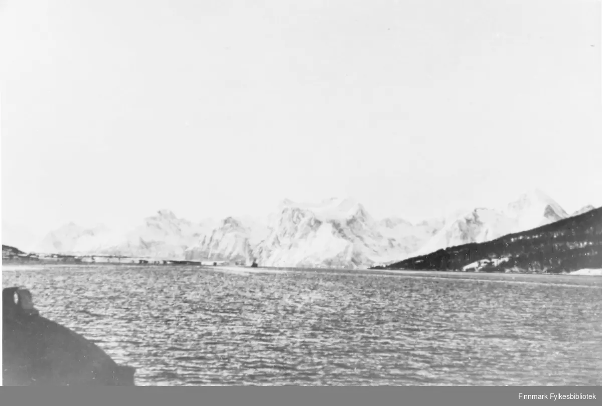 Reprofotografi av motiv tatt i Skjervøy, Troms. Sjøen ligger rolig i forgrunnen av bildet. Snekledde fjell ligger i bakgrunnen