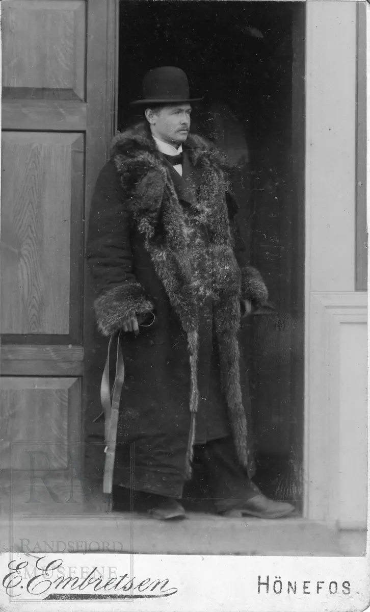 Mann stående i en døråpning, han har stor frakk med pelsbesetning og skalk (rund hatt).