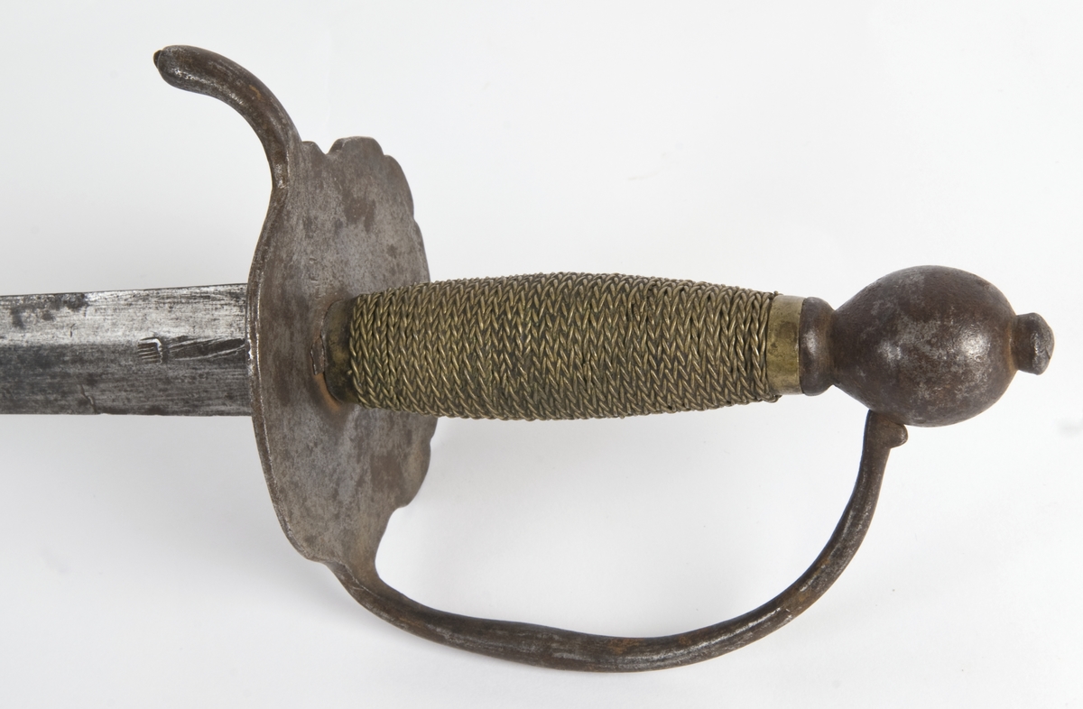 Dansk/Norsk Soldatkårde modell 1701. Plommeformet knapp med nitteknapp. Tregrep med messingtråds bevikling og hylse oppe og nede. Nyreformet parerplate med 5 fliker. Nedbøyd bakre parerstang. Fremre parerstang går over i håndbøylen og er tappet i knappen.
Rett tveegget klinge med 6 kantet tverrsnitt. Rester av etset dekor.