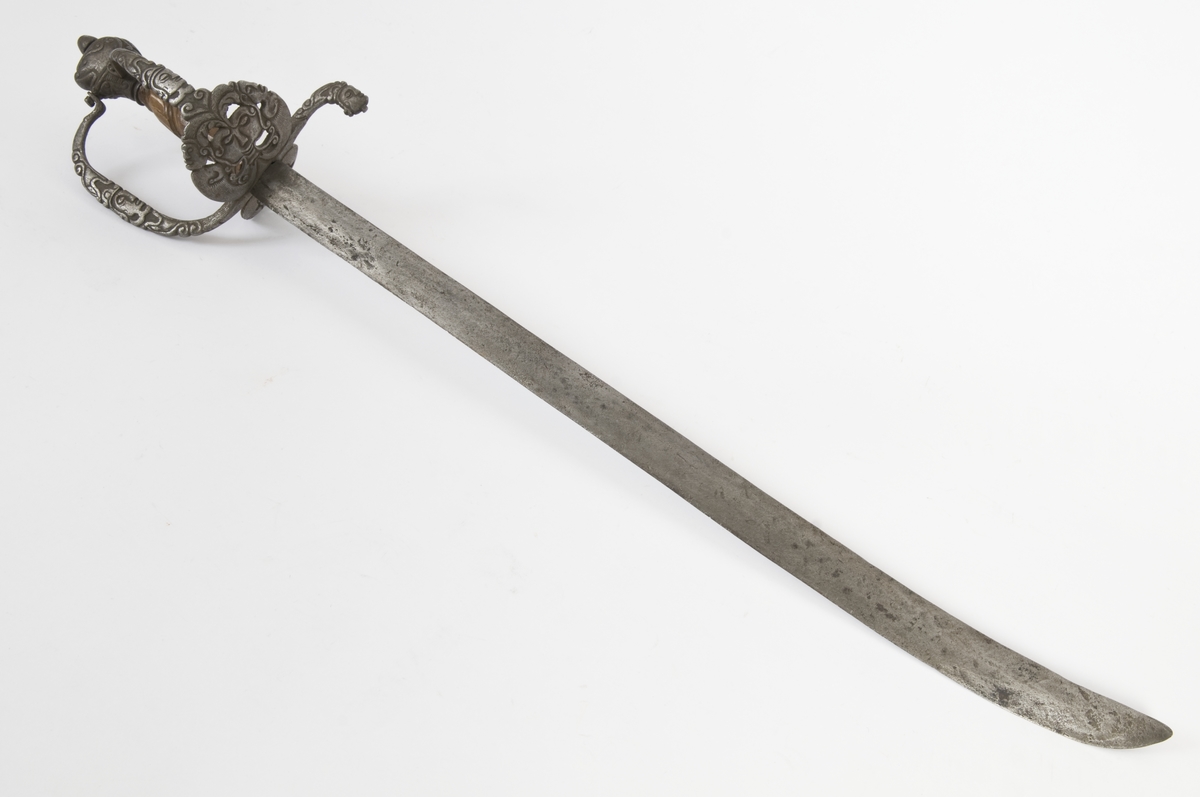 Hundehodeformet knapp med nitteknapp og skåret dekor.Tregrep  S formede parerstenger med fortykninger på endene.Fremre parerstang går over i håndbøylen, som er skrudd til knappen.Rund perforet stikkplate på utsiden med sidebøyle som er skrudd til knappen.Midere perforert stikkplate på innsiden med tombøyle. Hele festet har rikt skåret dekor bl a med masker.
Eneegget, krum klinge med kileformet tverrsnitt og ryggslipning mot spissen..