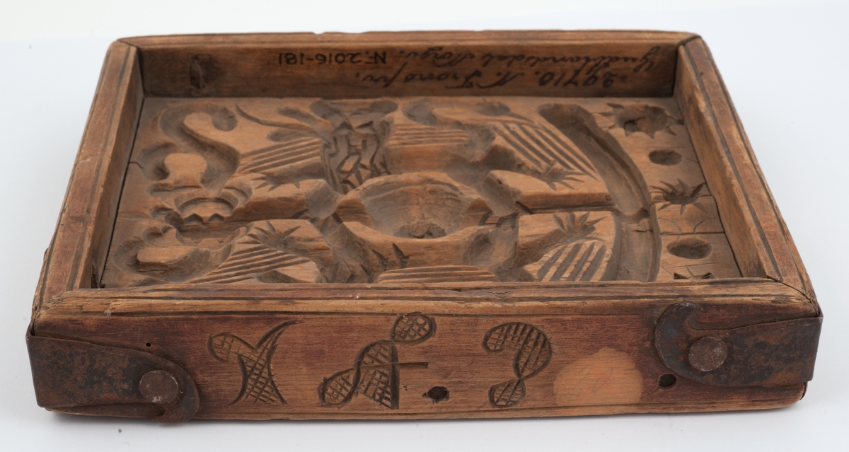 Firkantet kakeform med jernbeslag på hjørnene. Den ene siden av bunnen skåret med sol, måne, fabeldyr og et tre. Undersiden har blomster og rankedekor med 2 fugler. Litt skåret dekor på den andre siden.
Innskrift på den ene langsiden.