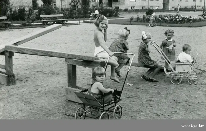 Barn på dumphuske og sittende i barnevogn ved en grusbelagt lekeplass. Blokkbebyggelse i bakgrunnen med reklameskilt for Monark Velosiped (Veltepetter) over inngangsparti.